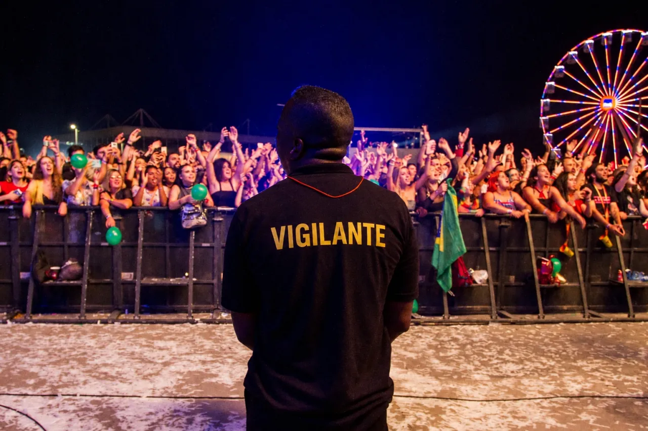 Vigilante acompanha o público diante de uma barreira durante um show noturno, em fotografia institucional sobre segurança de eventos.